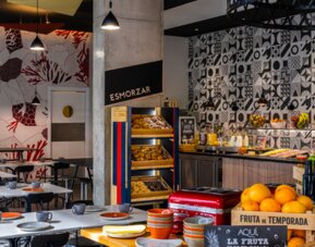 Breakfast buffet with pastries, fruit, cold foods, and drinks beside a patterned dining area at Hotel ibis Styles Barcelona City Bogatell.