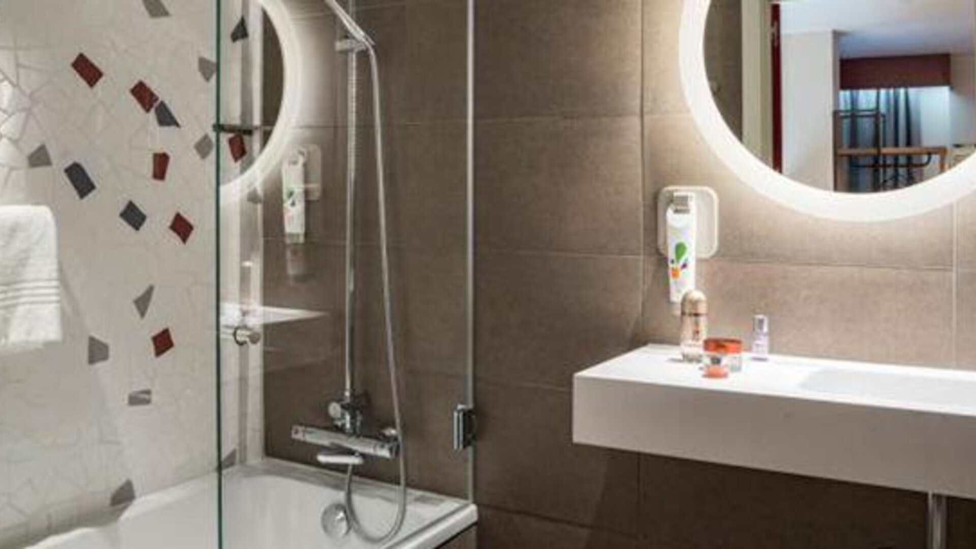 Bathroom with a glass-enclosed shower, white sink, round illuminated mirror, and tiled walls at Hotel ibis Styles Barcelona City Bogatell.