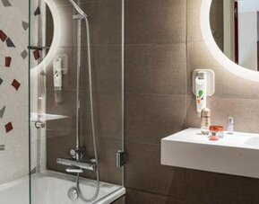 Bathroom with a glass-enclosed shower, white sink, round illuminated mirror, and tiled walls at Hotel ibis Styles Barcelona City Bogatell.
