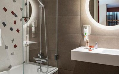 Bathroom with a glass-enclosed shower, white sink, round illuminated mirror, and tiled walls at Hotel ibis Styles Barcelona City Bogatell.