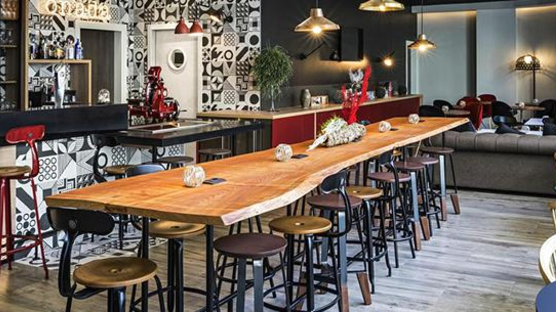 Hotel lounge and bar with a long wooden communal table, stools, sofas, pendant lights, and patterned walls at Hotel ibis Styles Barcelona City Bogatell.