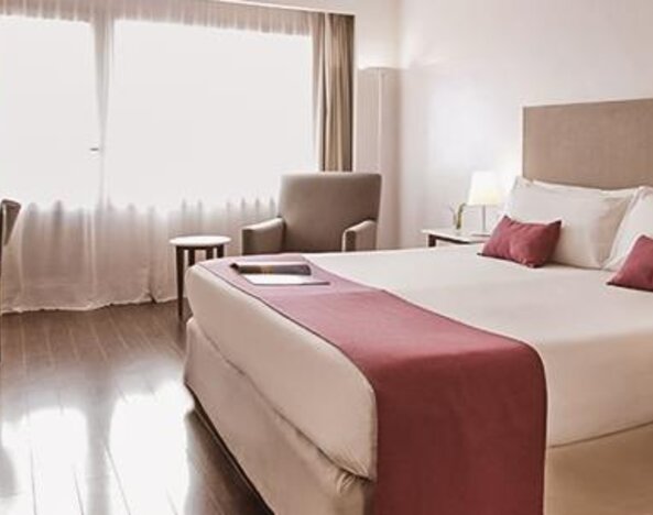 Guest room with a king bed, pink runner and pillows, armchair, desk, bedside tables, and large window at Dazzler by Wyndham Buenos Aires Recoleta.