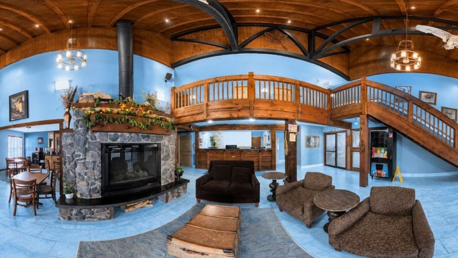 Spacious lodge-style hotel lobby and lounge featuring a central stone fireplace, upholstered seating, rustic wood ceiling, exposed beams, reception desk and sweeping wooden staircase.