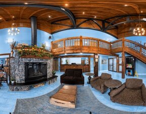 Spacious lodge-style hotel lobby and lounge featuring a central stone fireplace, upholstered seating, rustic wood ceiling, exposed beams, reception desk and sweeping wooden staircase.