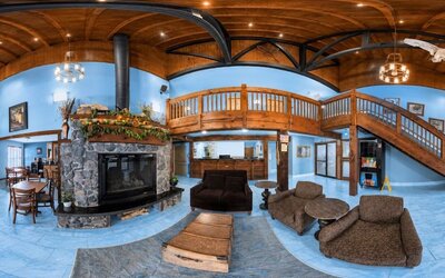Spacious lodge-style hotel lobby and lounge featuring a central stone fireplace, upholstered seating, rustic wood ceiling, exposed beams, reception desk and sweeping wooden staircase.