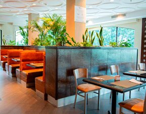 Hotel dining area with tables, chairs, orange booth seating and tropical plants beside large windows at Holiday Inn Panama Canal.