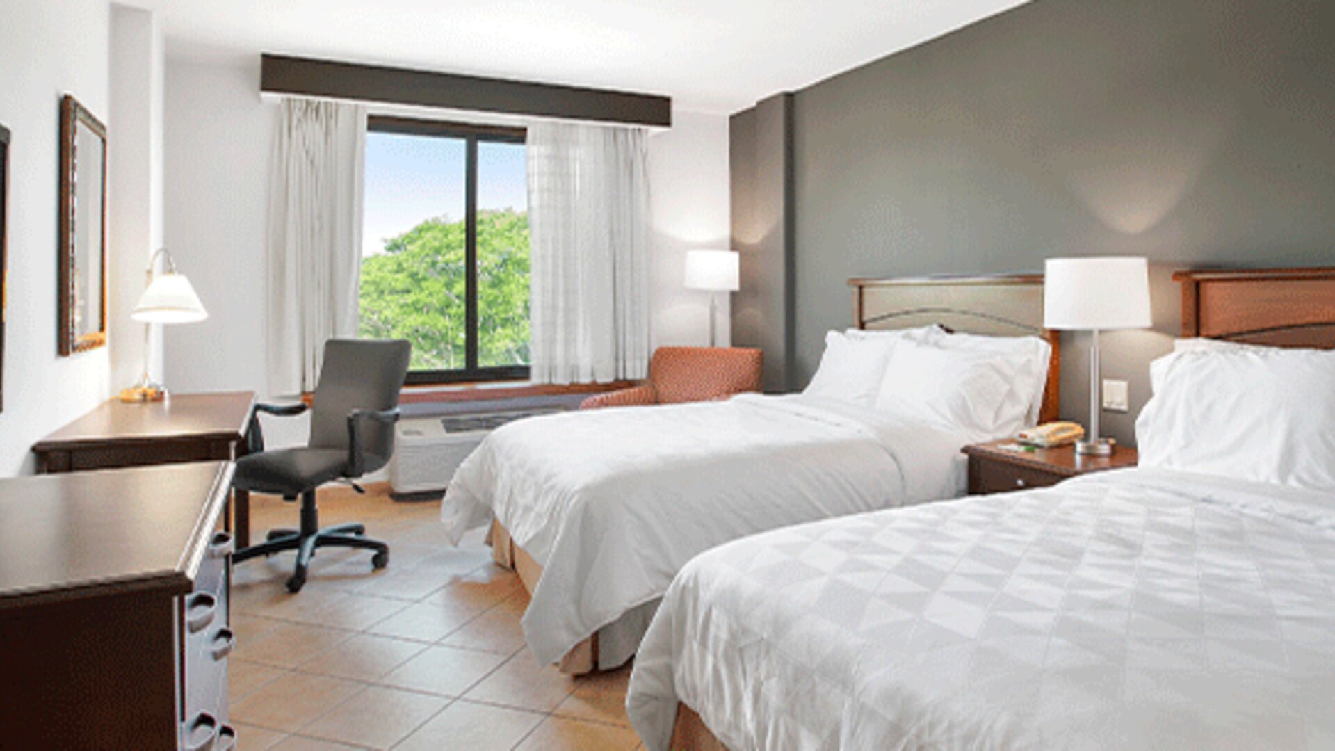 Guest room with two beds, bedside table, desk, office chair and large window overlooking greenery at Holiday Inn Panama Canal.