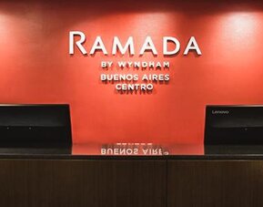 Hotel reception desk with two computer terminals and a red accent wall displaying the Ramada by Wyndham Buenos Aires Centro logo.