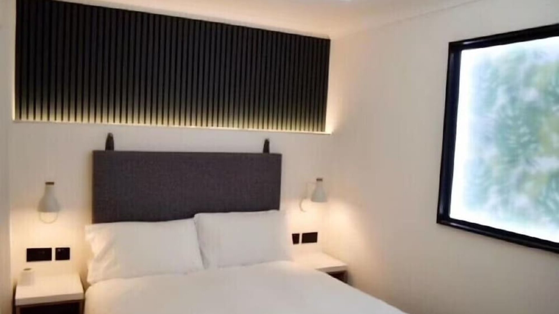 Double guest room with a white bed, dark upholstered headboard, bedside tables, wall-mounted reading lights and a large window at Victoria Inn.