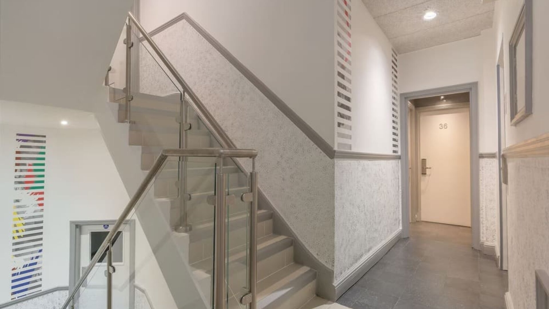 Interior staircase and hallway at Victoria Inn with glass stair railings, gray flooring, white walls and guest room doors.