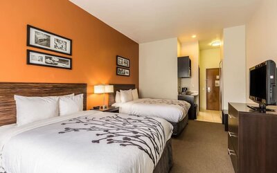 Double guest room with two beds, white bedding, orange accent wall, framed artwork, bedside lamp, television, desk, refrigerator, microwave, and coffee maker at Sleep Inn & Suites Stafford - Sugar Land.
