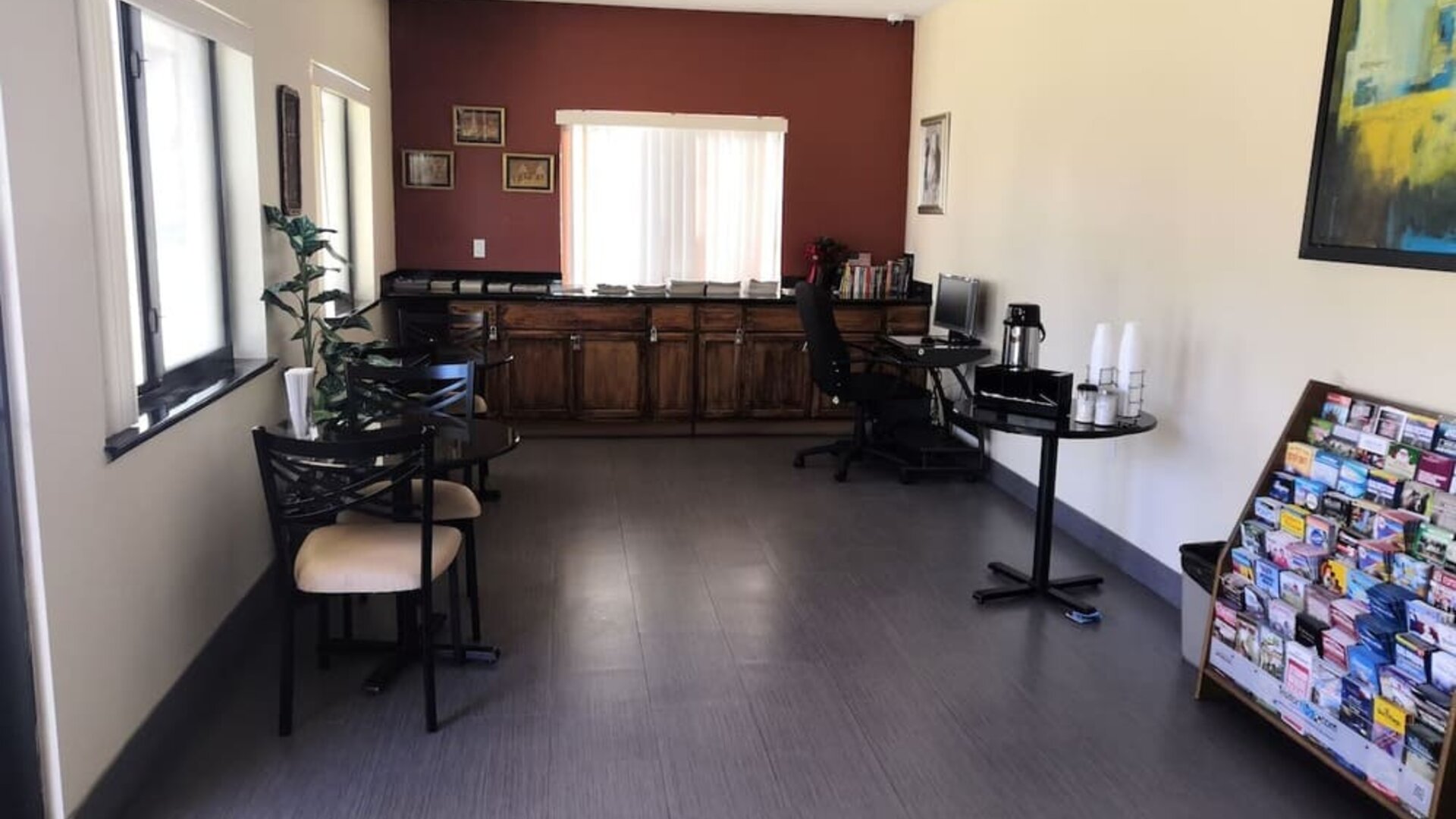Hotel common area with tables and chairs, a long wooden counter, work desk with computer, beverage station, windows, and a brochure rack at Scottish Inn & Suites.