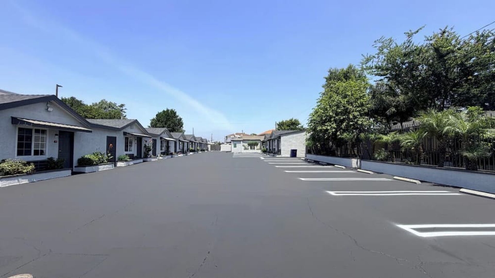 Wide daytime view of Scenic Motel’s spacious paved parking area surrounded by rows of single-story guest rooms with individual exterior entrances, white-lined parking spaces, landscaped greenery, trees, and palm plants beneath a clear blue sky.