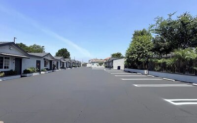 Wide daytime view of Scenic Motel’s spacious paved parking area surrounded by rows of single-story guest rooms with individual exterior entrances, white-lined parking spaces, landscaped greenery, trees, and palm plants beneath a clear blue sky.