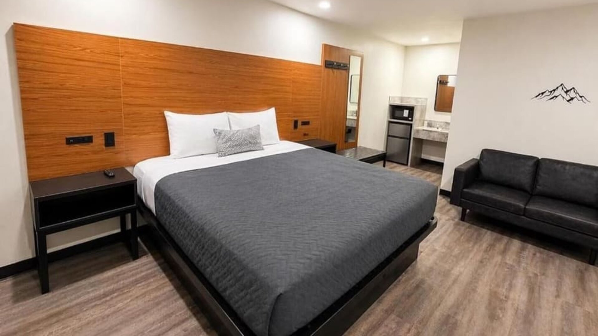 Modern standard king room with a large bed dressed in gray and white bedding, expansive wood-paneled headboard, black bedside tables, black loveseat, microwave, mini refrigerator, vanity area, recessed ceiling lights, wood-look flooring, and minimalist mo