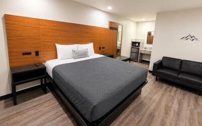 Modern standard king room with a large bed dressed in gray and white bedding, expansive wood-paneled headboard, black bedside tables, black loveseat, microwave, mini refrigerator, vanity area, recessed ceiling lights, wood-look flooring, and minimalist mo