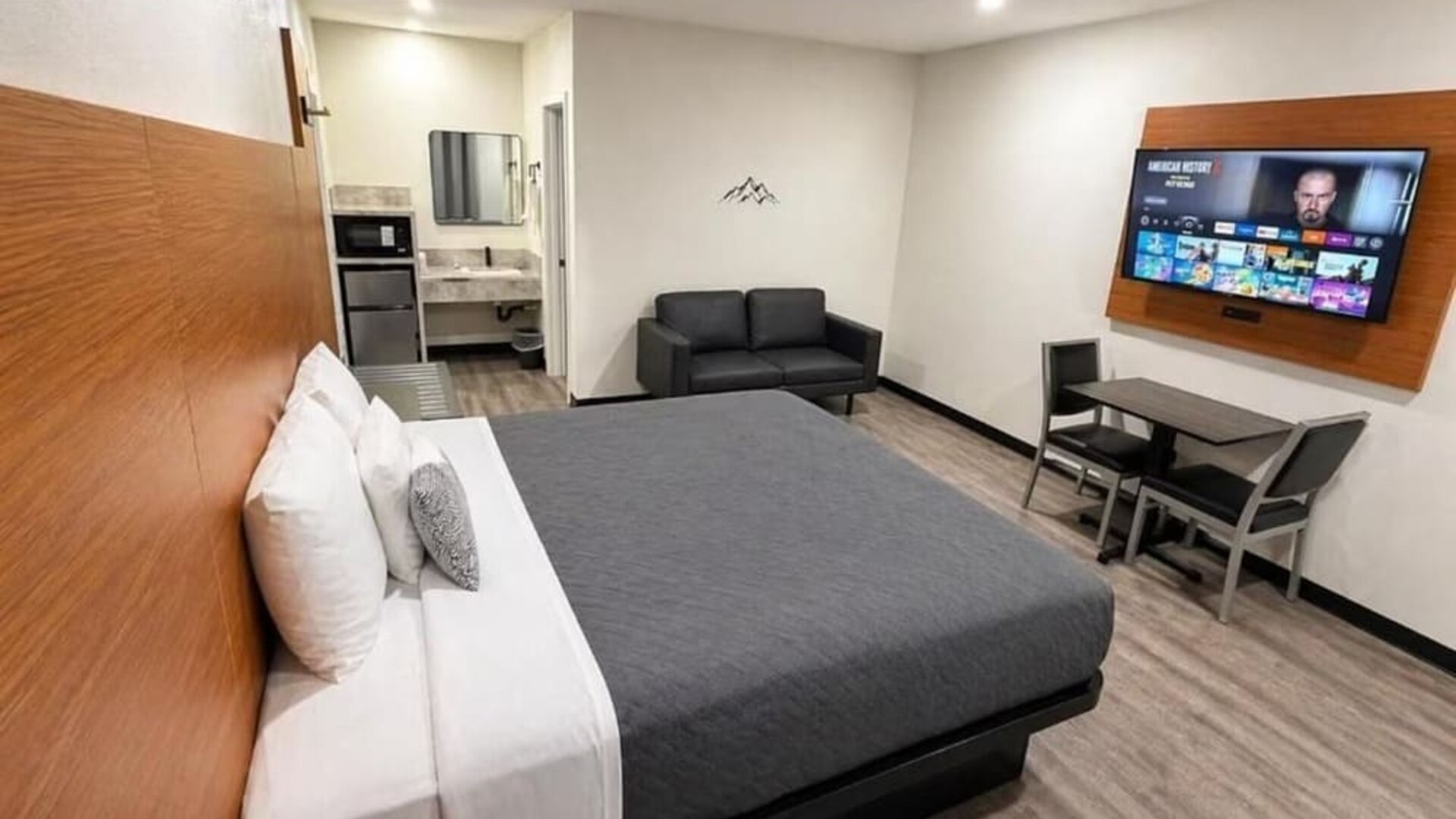 Spacious modern king room with a large bed dressed in gray and white linens, wood-paneled headboard, black loveseat, wall-mounted flat-screen TV, dining table with two chairs, microwave, mini refrigerator, vanity area, recessed lighting, and wood-look flo