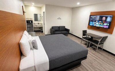 Spacious modern king room with a large bed dressed in gray and white linens, wood-paneled headboard, black loveseat, wall-mounted flat-screen TV, dining table with two chairs, microwave, mini refrigerator, vanity area, recessed lighting, and wood-look flo
