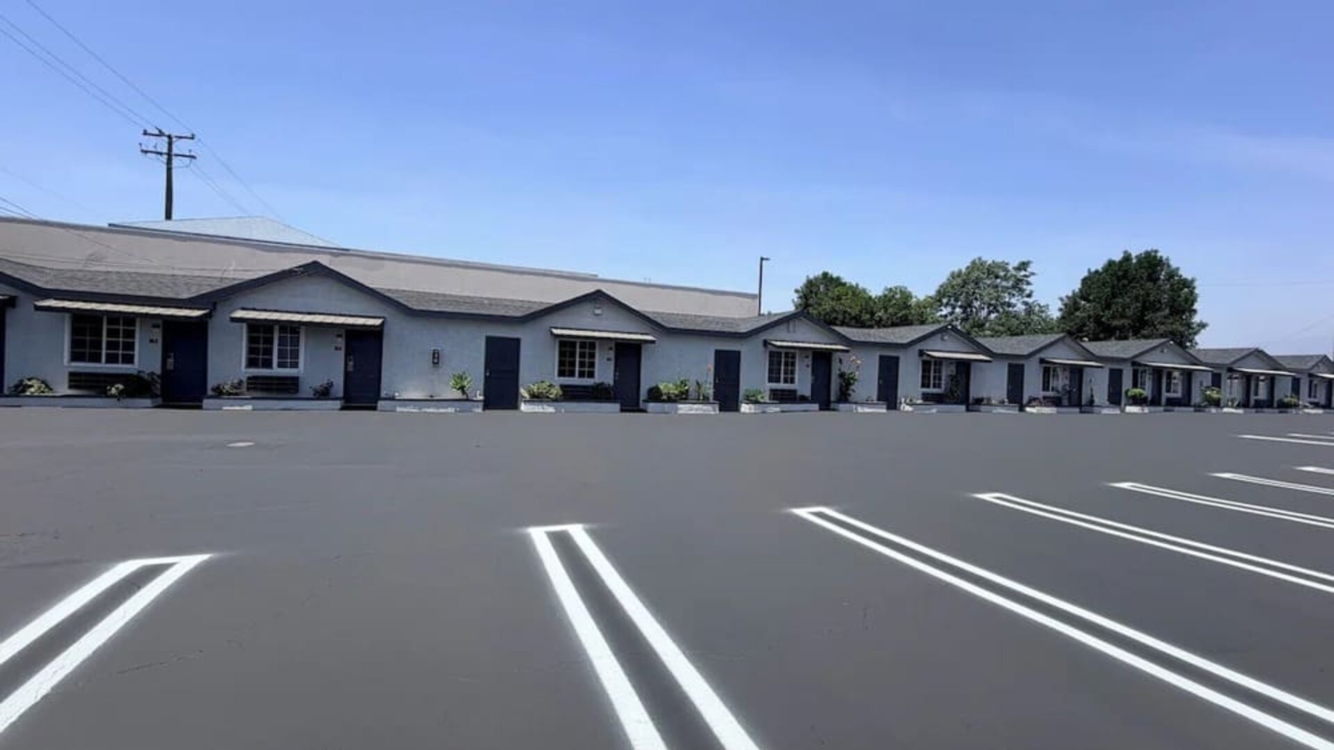 Daytime view of the Scenic Motel parking area with a clearly marked accessible parking space in the foreground and rows of single-story guest rooms with individual entrances, windows, small awnings, landscaped shrubs, and parking spaces extending into the