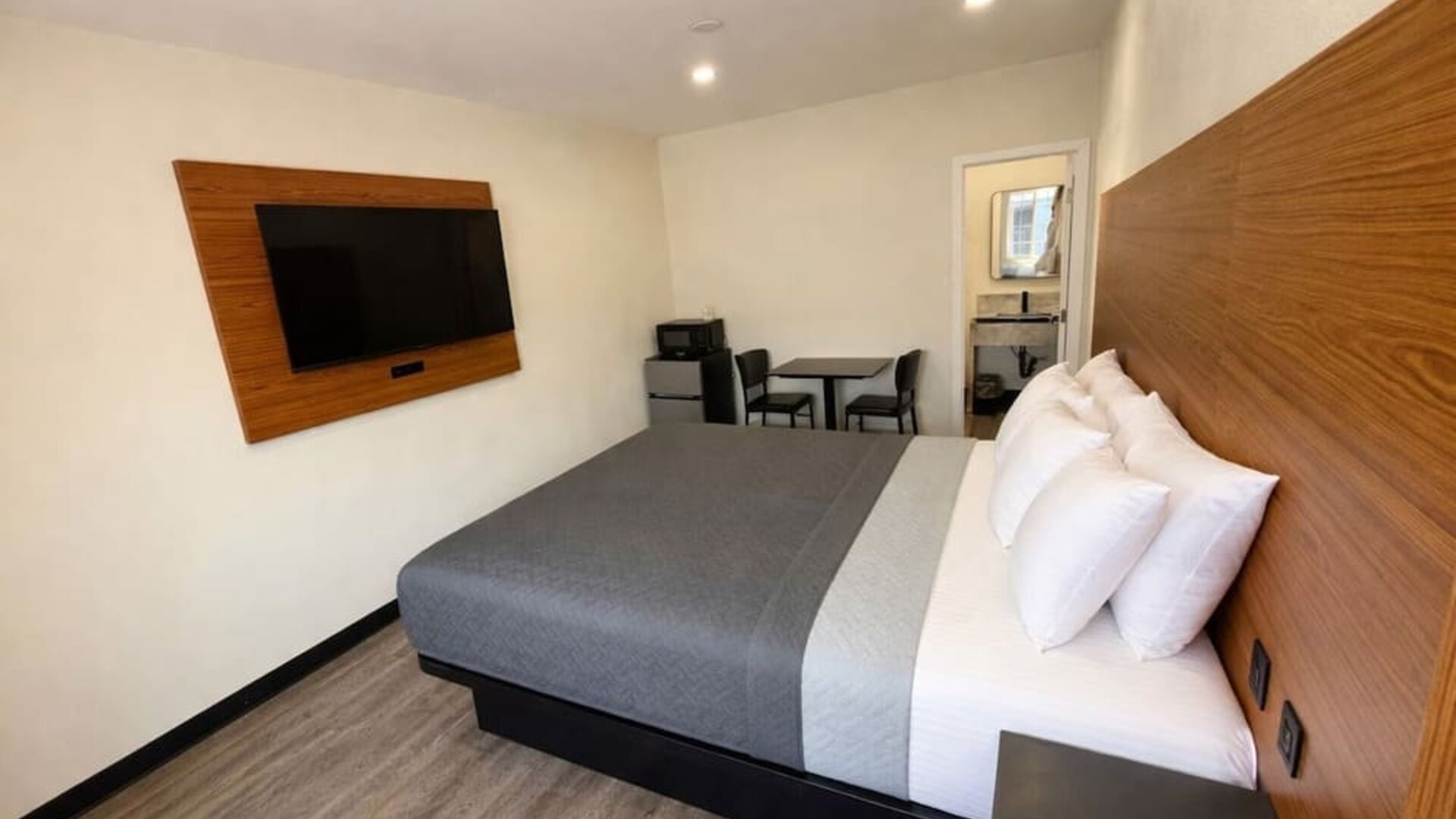 Bright modern guest room with a king-size bed dressed in gray and white bedding, a large wood-paneled headboard, wall-mounted flat-screen TV, small dining table with two chairs, microwave, mini refrigerator, wood-look flooring, and an open doorway leading