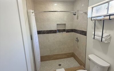 Modern private bathroom featuring a spacious tiled walk-in shower with a white curtain, built-in wall niche and showerhead, alongside a white toilet, wall-mounted towel rack with folded towels, frosted window, recessed lighting, and gray wood-look floorin