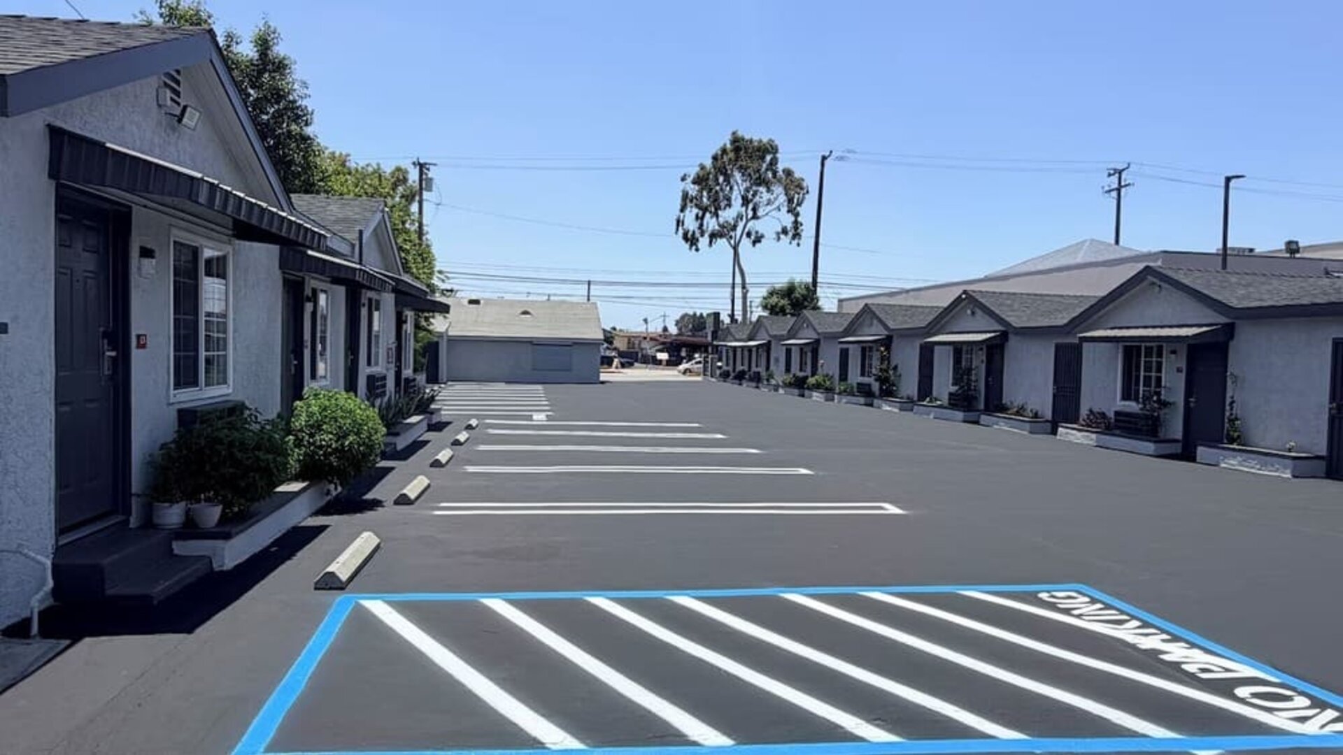 Wide exterior view of Scenic Motel showing a long row of single-story guest rooms with individual doors, windows, small covered awnings, and landscaped planters facing a large freshly paved parking area with clearly marked spaces under a bright blue sky.