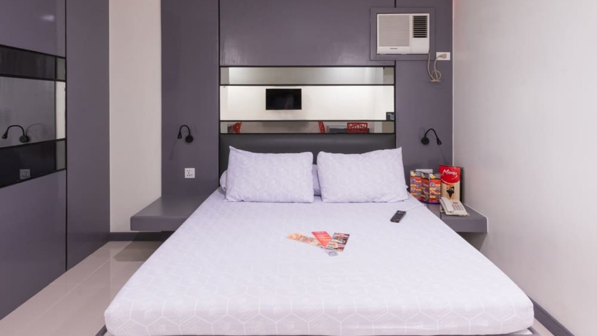 Modern double room with a white bed, purple-gray built-in headboard and wall cabinetry, mirrored panels, bedside shelves, wall-mounted reading lights, air-conditioning unit, telephone, and informational brochures at Hotel Sogo Guadalupe.