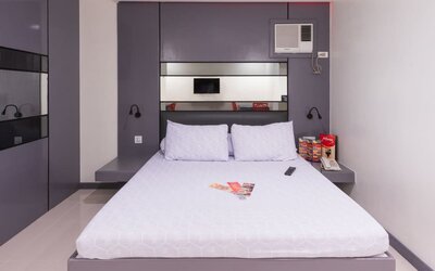 Modern double room with a white bed, purple-gray built-in headboard and wall cabinetry, mirrored panels, bedside shelves, wall-mounted reading lights, air-conditioning unit, telephone, and informational brochures at Hotel Sogo Guadalupe.