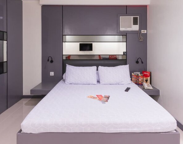 Modern double room with a white bed, purple-gray built-in headboard and wall cabinetry, mirrored panels, bedside shelves, wall-mounted reading lights, air-conditioning unit, telephone, and informational brochures at Hotel Sogo Guadalupe.