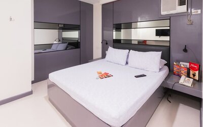 Modern double room with a white bed, purple-gray built-in headboard and mirrored panels, bedside shelves, reading lights, air-conditioning unit, telephone, and brochures at Hotel Sogo Guadalupe.