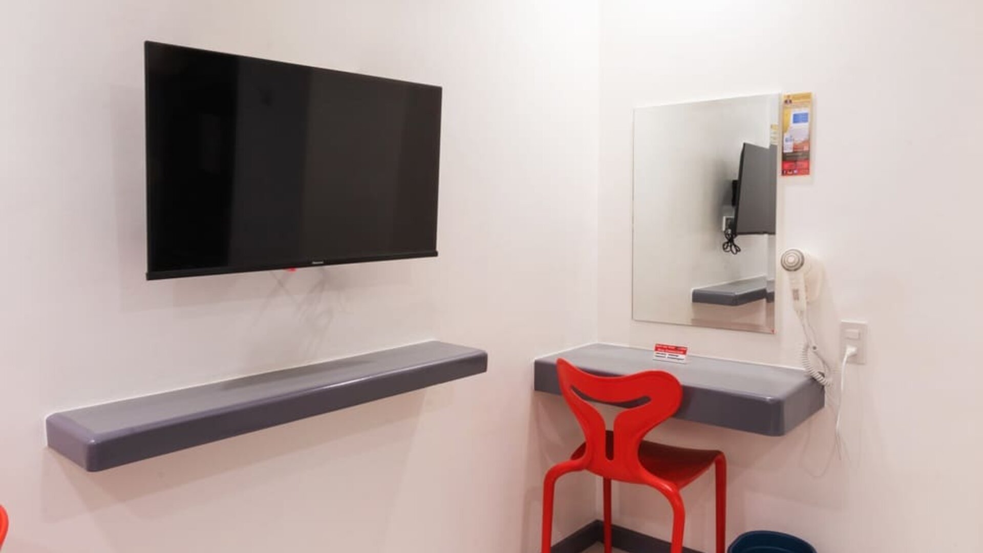 In-room workspace with a wall-mounted flat-screen TV, floating gray desk shelves, rectangular mirror, red chair, wall-mounted hairdryer, and waste bin at Hotel Sogo Guadalupe.