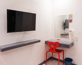 In-room workspace with a wall-mounted flat-screen TV, floating gray desk shelves, rectangular mirror, red chair, wall-mounted hairdryer, and waste bin at Hotel Sogo Guadalupe.