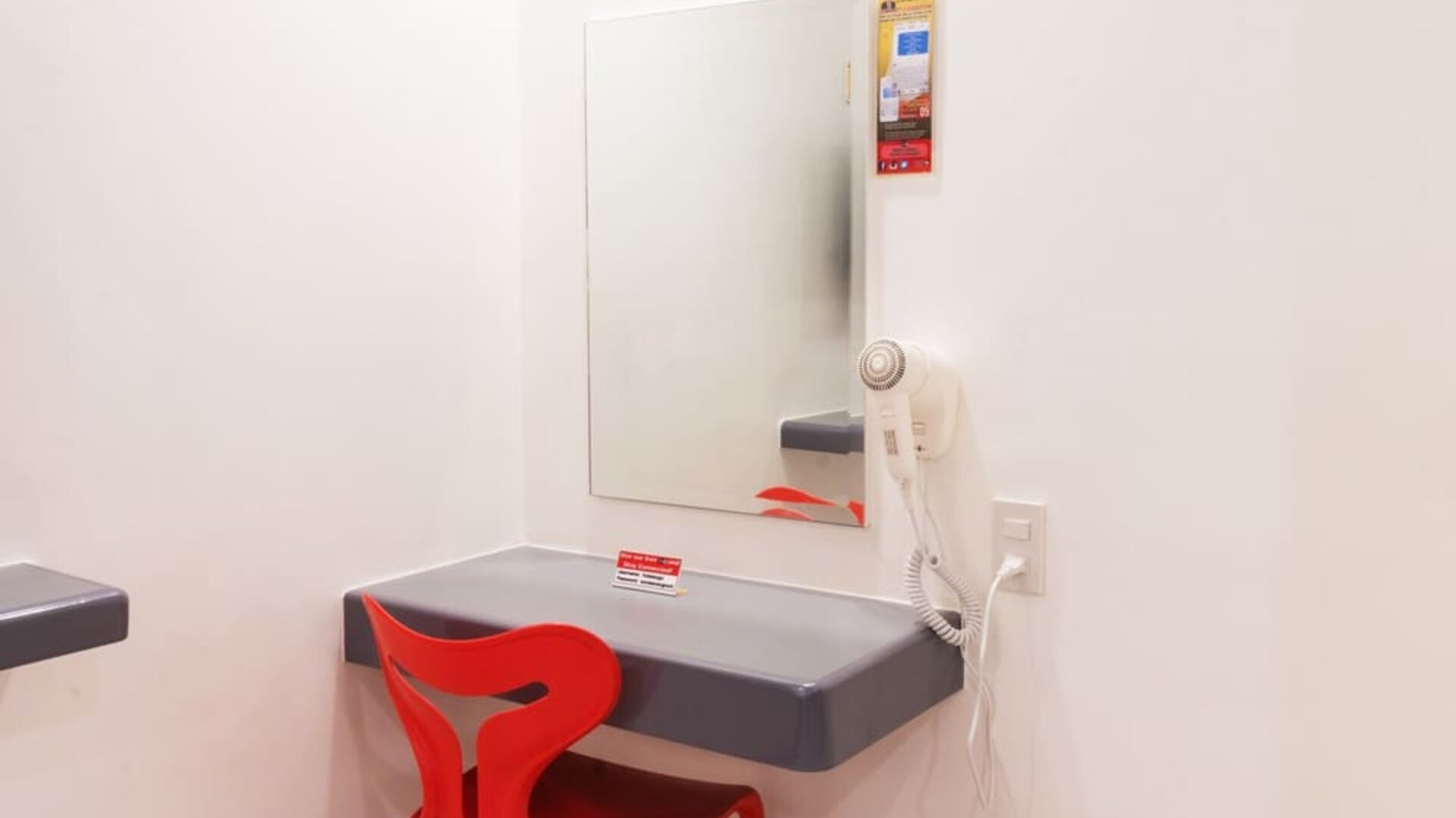 In-room workspace featuring a floating gray desk, rectangular wall mirror, bright red chair, wall-mounted hairdryer, and electrical outlet at Hotel Sogo Guadalupe.
