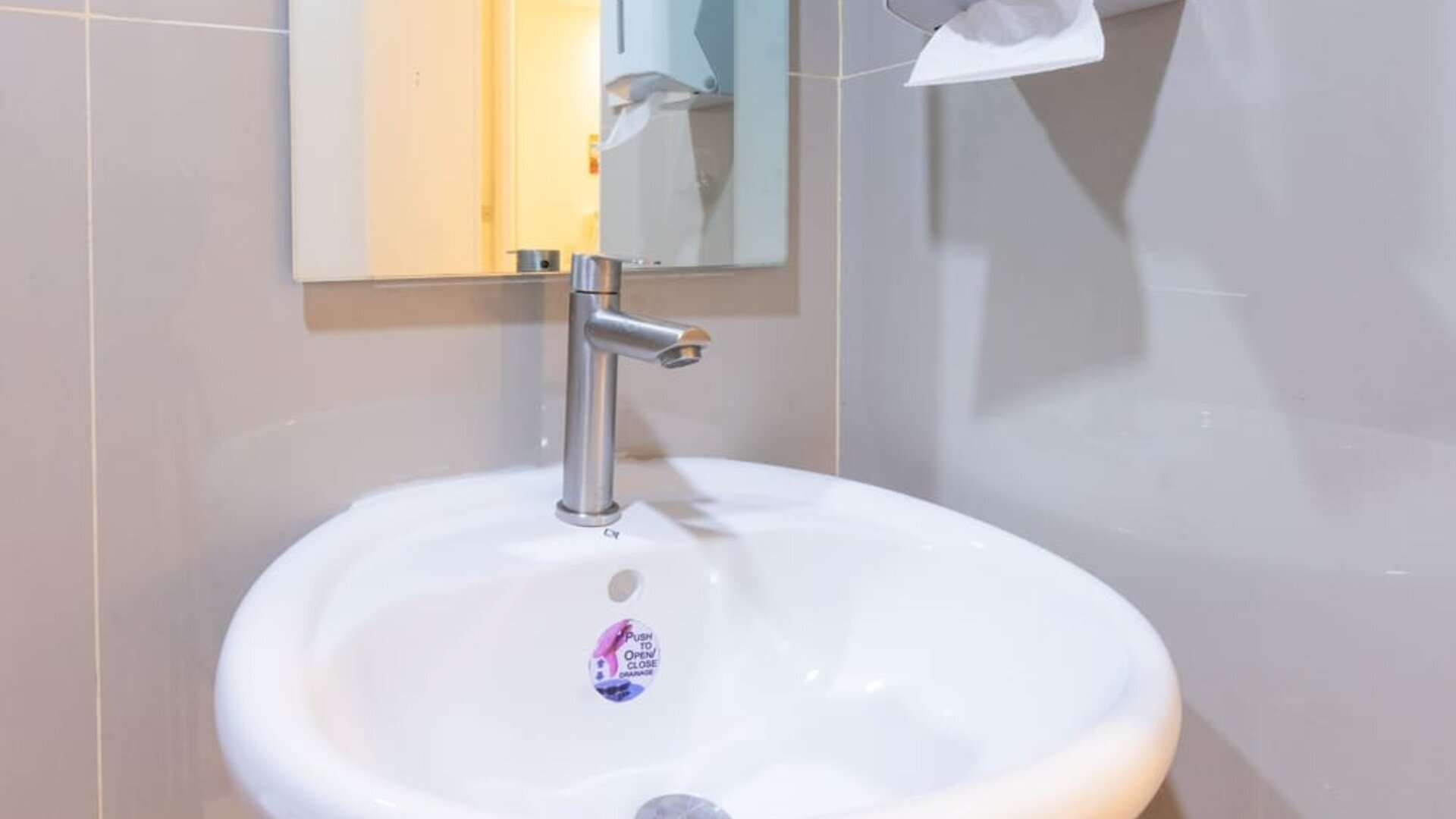 Close-up of a white round bathroom sink with a modern silver faucet, rectangular wall mirror, and wall-mounted soap and paper towel dispensers at Hotel Sogo Guadalupe.