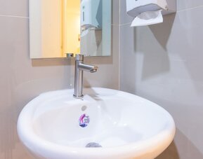 Close-up of a white round bathroom sink with a modern silver faucet, rectangular wall mirror, and wall-mounted soap and paper towel dispensers at Hotel Sogo Guadalupe.