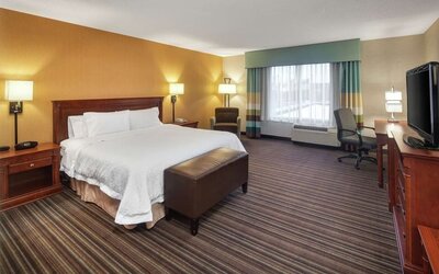 Spacious hotel room with a king-size bed, white bedding, bedside tables, armchair, work desk and chair, large window, wooden dresser, and flat-screen TV.