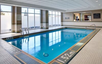 Bright indoor hotel swimming pool surrounded by tiled flooring, large windows, pool ladders, and an accessible handrail.