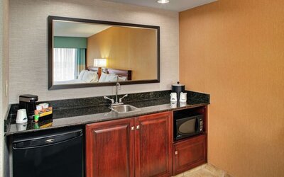 Hotel room kitchenette with granite countertop, sink, mini refrigerator, microwave, coffee maker, storage cabinets, cups, and beverage supplies.