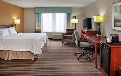 Spacious king guest room with white bedding, work desk and ergonomic chair, armchair, flat-screen TV, mini refrigerator, coffee maker, and a large window.