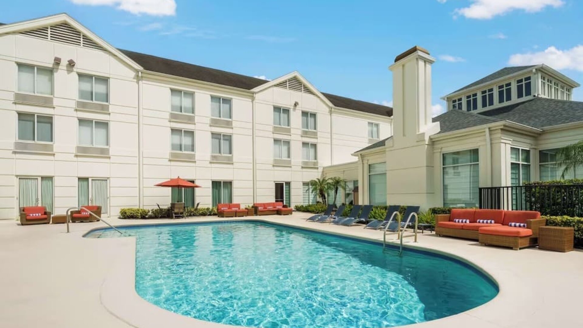 Curved outdoor hotel swimming pool with lounge chairs, orange cushioned seating, an umbrella and a bright three-story hotel building surrounding the courtyard.