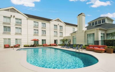 Curved outdoor hotel swimming pool with lounge chairs, orange cushioned seating, an umbrella and a bright three-story hotel building surrounding the courtyard.