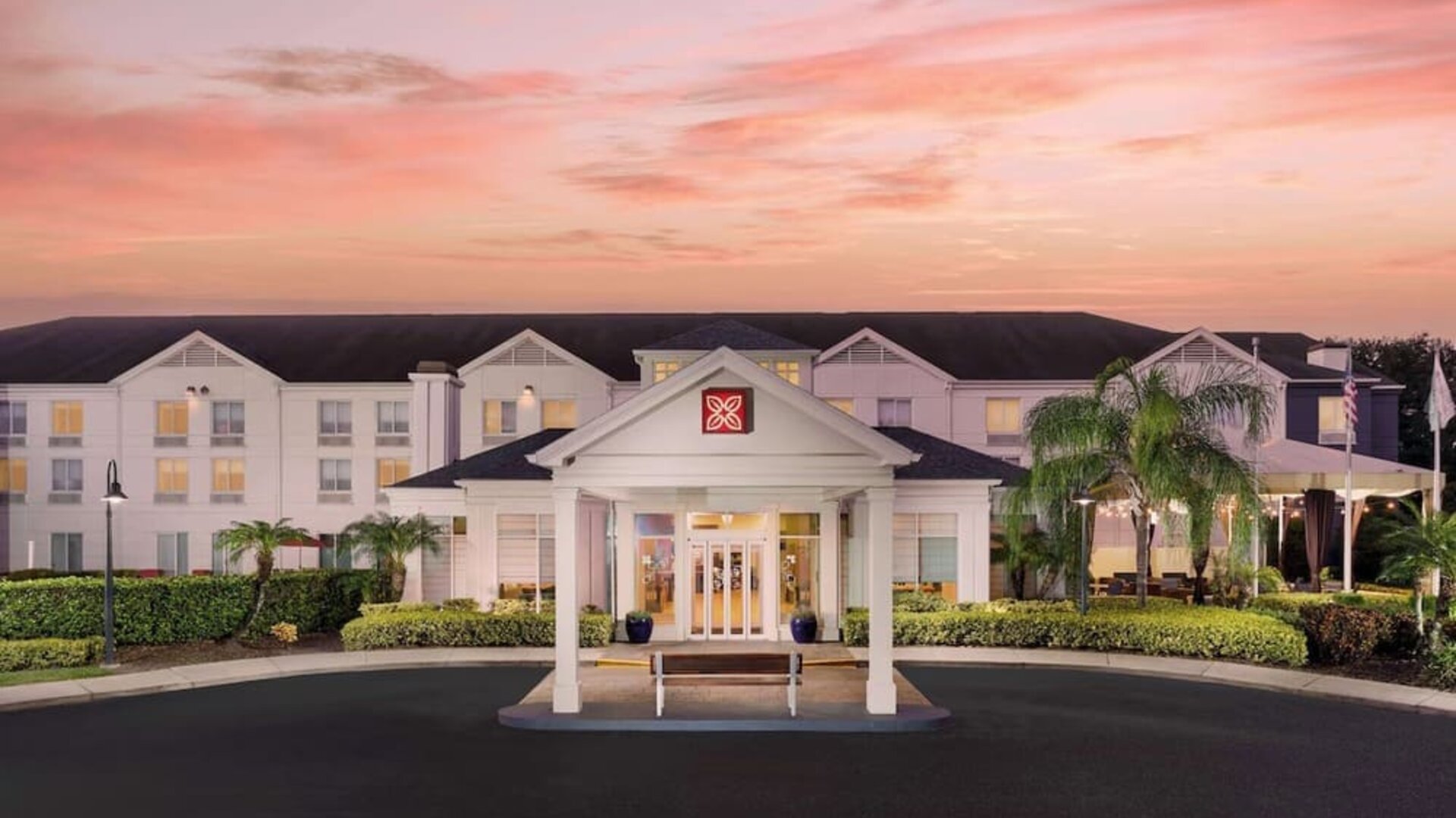 Front exterior of the hotel at sunset with a covered entrance, white three-story façade, palm trees, landscaped grounds and a pink-orange evening sky.