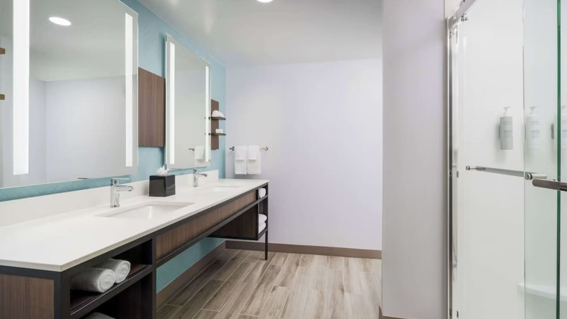 Spacious modern hotel bathroom with a long double-sink vanity, illuminated mirrors, open towel storage and a glass-enclosed walk-in shower.