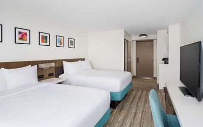 Spacious hotel guest room with two white beds on turquoise bases, colorful framed artwork, large flat-screen TV, teal desk chair and modern wood-look flooring.`