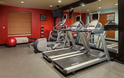 Hotel fitness center with two treadmills, elliptical machine, exercise bike, dumbbell rack, weight bench, stability ball, wall-mounted TV and mirrored wall.
