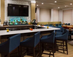 Stylish hotel bar with a white countertop, blue upholstered bar stools, illuminated liquor display, beer taps, large TV and adjacent dining tables.