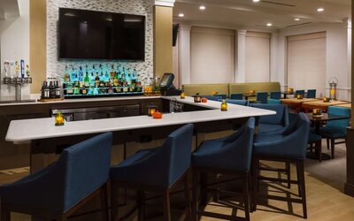 Stylish hotel bar with a white countertop, blue upholstered bar stools, illuminated liquor display, beer taps, large TV and adjacent dining tables.