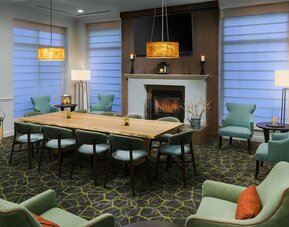 Cozy hotel lounge with a central fireplace, large communal wood table, mint-green armchairs, floor lamps and warm pendant lighting.