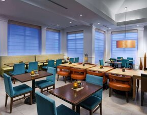 Contemporary hotel dining area with turquoise chairs, cushioned booth seating, dark wood tables, warm pendant lighting and large windows with privacy shades.