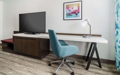 Hotel guest room workspace with a long white desk, teal rolling chair, desk lamp, large flat-screen TV, dark wood cabinet and colorful framed artwork.